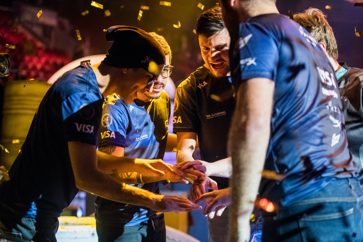 Esports teammates in matching uniforms share a joyful moment, celebrating with a group huddle under a shower of golden confetti after a victory.